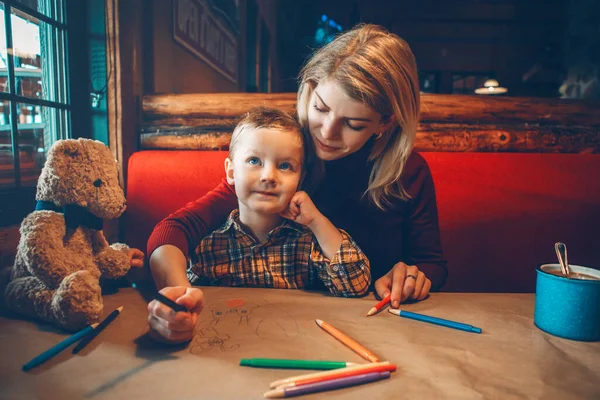 Young Caucasian mother and son sitting in restaurant drawing with colored pencils on craft brown paper. Family together in cafe. Lifestyle authentic real people. Mothers Day lifestyle.