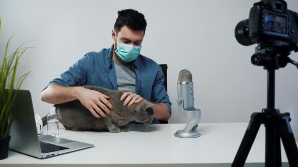 Αρσενικό blogger streaming φορώντας ιατρική μάσκα Παίζοντας με γάτα. Δημιουργός περιεχομένου βίντεο. Βίντεο Ιού Concept. Blogger Ιατρική Μάσκα Χρήση. Ο άνθρωπος παίζει με το κατοικίδιο ζώο — Αρχείο Βίντεο
