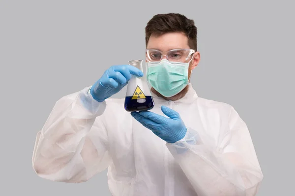 Manlig laboratoriearbetare i kemisk kostym, bär medicinsk mask och glasögon titta på kolv med blå flytande strålning tecken. Vetenskap, medicin, viruskoncept. Blå vätska — Stockfoto