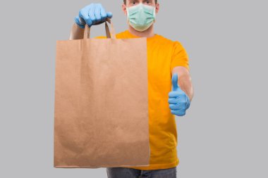 Ellerinde kağıt torbayla tıbbi maske ve eldiven takan teslimatçı. Sarı Tişört Teslimatçısı. Ev yemeği teslimatı. İzole edilmiş