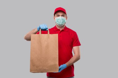 Ellerinde kağıt torbayla tıbbi maske ve eldiven takan teslimatçı. Kırmızı üniformalı teslimatçı çocuk. Ev yemeği teslimatı. Kağıt Torba. İzole edilmiş