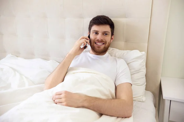 Ο άντρας που μιλάει στο τηλέφωνο στο κρεβάτι. Πρωί στο σπίτι. Ψύξη άνθρωπος χρησιμοποιώντας το τηλέφωνο στο κρεβάτι. Καραντίνα, κατ 'οίκον εργασία, ιατρική περίθαλψη. — Φωτογραφία Αρχείου