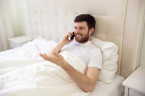 Man Talking on Phone in Bed. Morning at Home. Smilling Man Using Phone in Bed. Quarantine, Home Work, Medical Care.