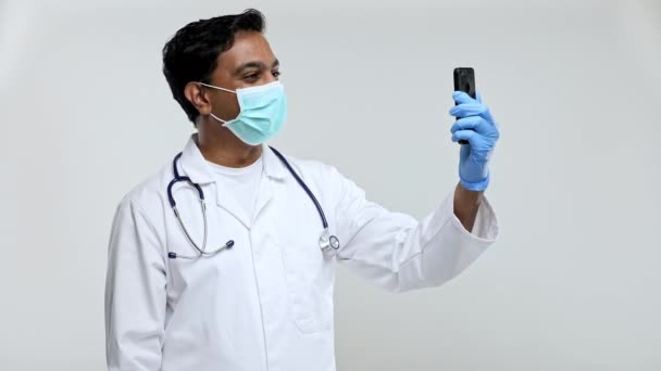 Médico teniendo videollamada en el teléfono usando máscara médica y guantes aislados. Hombre indio Doctor Video Call. Medicina en línea. Médico usando teléfono . — Vídeos de Stock