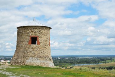 Soldaki yeşil çimenlikte bulutlu bir gökyüzüne bakan antik taş kule.