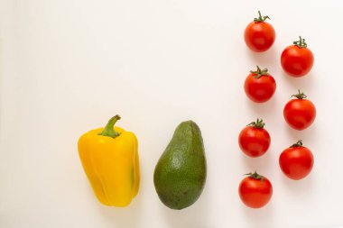 Taze kırmızı kiraz domatesleri dikey bir çizgide, beyaz arka planda izole edilmiş sarı dolma biber ve avokado eşliğinde duruyor.