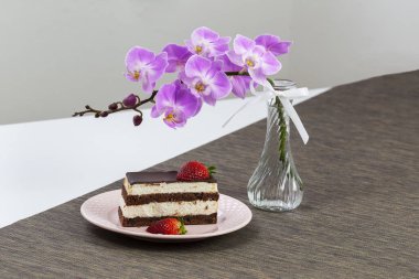 Tatlı - Vanilya dolgulu çikolatalı pasta üzerine beyaz çikolata jölesi ve beyaz bir tabağa beyaz çikolata serpiştirilmiş. Arkaplanda orkideli pembe vazo.
