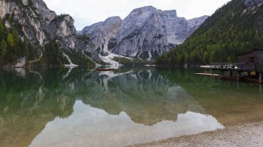 İtalyan Dolomitler, Lago di Braiescalm, yüksek kıyıları ağaçlarla kaplı mavi-yeşil göl yüzeyi. Soldaki ahşap korkulukta, sudaki tahta ayaklıkların üzerinde duran eve doğru gidiyor..