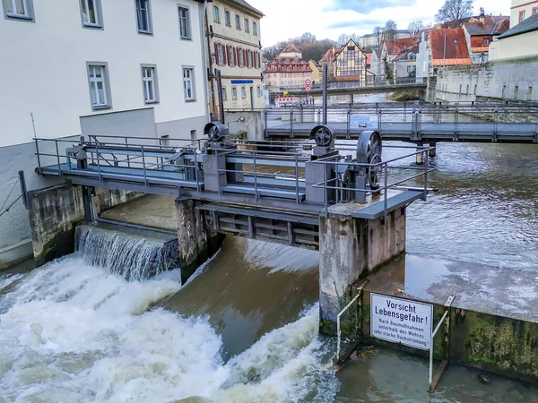 A dam and a clearing station for water on a river in the center of a European city with dirty water and beautiful houses in the background