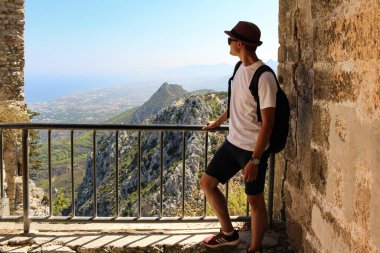 Sırt çantası ve şapkası olan genç bir turist balkondan dağın tepesine, mavi denize ve berrak gökyüzüne bakıyor. Kıbrıs 'ta turizm ve dinlenme.