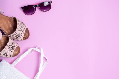 Pembe arka planda yaz aksesuarları bulunmaktadır: sandaletler, güneş gözlükleri ve bir plaj çantası.