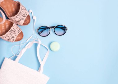 Mavi arka planda yaz aksesuarları bulunmaktadır: müzik kulaklıkları, plaj çantası, güneş gözlükleri ve dudak kremi ve sandaletler.