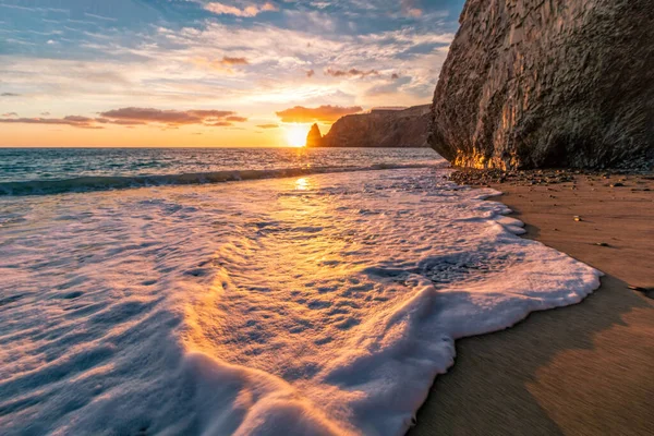 Bright sea sunset. The waves crash into the rock, lit by the warm sunset, sand and pebbles, volcanic basalt as in Iceland. Sea wave breaks into splashes and white foam. Never-ending beauty of nature