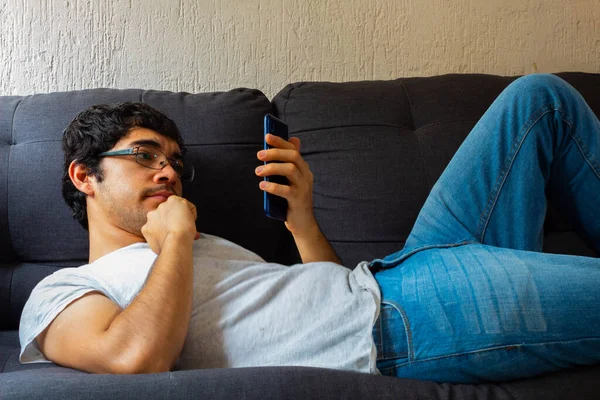 Hispanic young man with clear brown skin wearing glasses using smarthphone on a couch