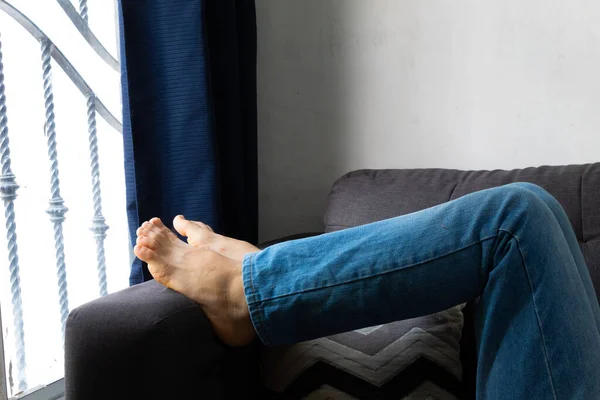 Legs of a man in blue jeans with bare feet resting and relaxing on a sofa receiving light from a window