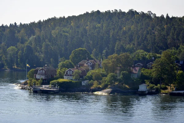 Baltık Denizi, İsveç kıyısı. 25 Temmuz 2014. Dar bir kıyıda beyaz pencereli geleneksel köy kahverengi evleri ve arka bahçelerinde yeşil hastalıklar var..