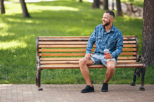 A guy with lemonade is resting in a park on a bench in the spring. Sunny day. Green foliage.