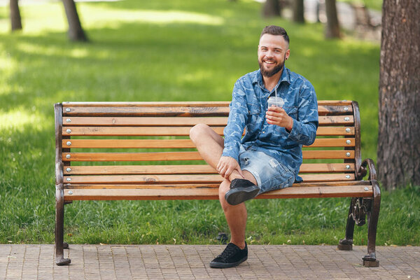 A guy with lemonade is resting in a park on a bench in the spring. Sunny day. Green foliage.
