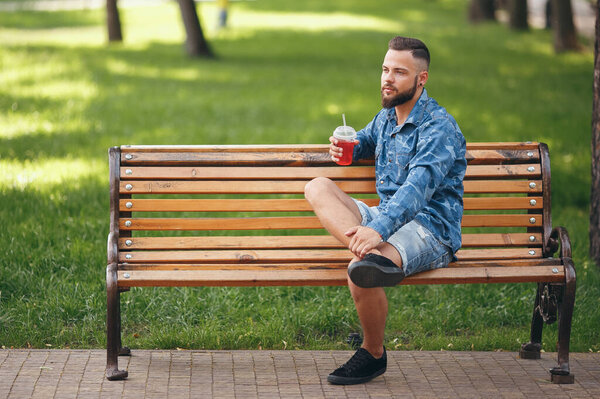 A guy with lemonade is resting in a park on a bench in the spring. Sunny day. Green foliage.