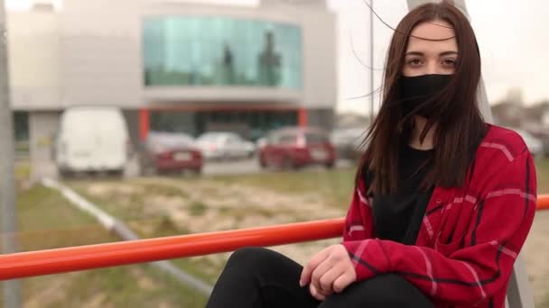 医療用マスクの少女が通りのベンチに腰を下ろしている。路上のマスクの少女の肖像画。コロナウイルスパンデミックの概念。通りにマスク. — ストック動画