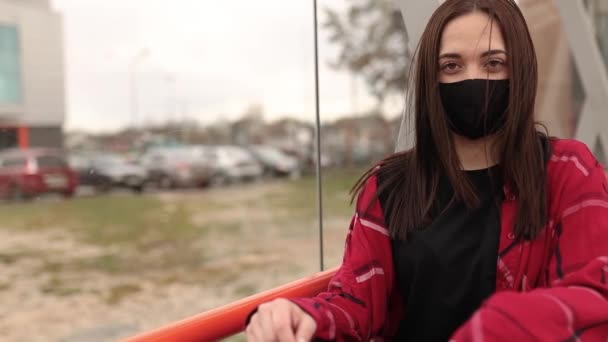 Meisje met een medisch masker zit op een bankje op straat. Portret van een meisje met een masker op straat. Coronavirus pandemie concept. Masker op straat. — Stockvideo