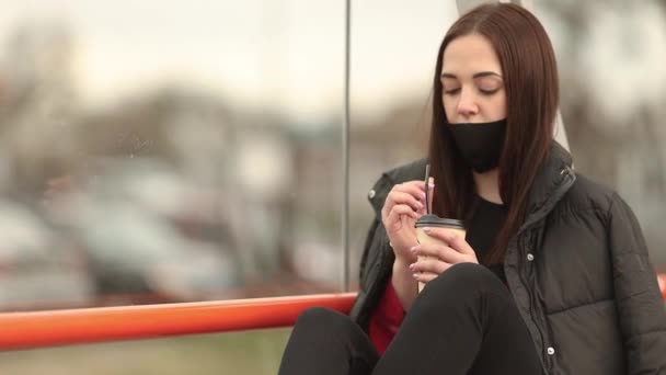 Uma menina com uma máscara médica senta-se em um banco na rua e bebe café. Retrato de uma menina com uma máscara na rua do café. Conceito de pandemia de coronavírus. Máscara na rua . — Vídeo de Stock