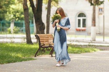 Güneşli bir bahar parkında elbise giymiş şık bir kız. Güzel bir kızın sakin portresi. Baharda elinde bir buketle duruyor.