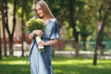Güneşli bir bahar parkında elbise giymiş şık bir kız. Güzel bir kızın sakin portresi. Baharda elinde bir buketle duruyor.