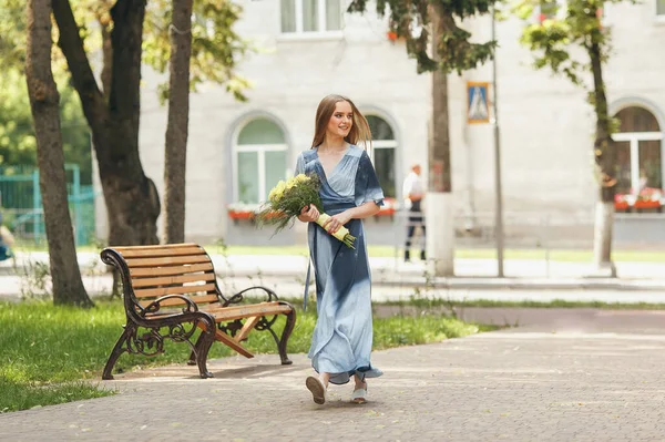 Fille élégante posant dans une robe dans un parc printanier ensoleillé. Portrait calme d'une belle fille debout avec un bouquet au printemps — Photo