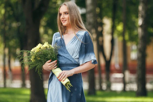 Fille élégante posant dans une robe dans un parc printanier ensoleillé. Portrait calme d'une belle fille debout avec un bouquet au printemps — Photo