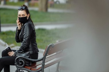 Siyah koruyucu maskeli esmer kadın portresi güneşli bir bahar gününde parkta bankta oturuyor. Cep telefonuyla konuşan kadın.