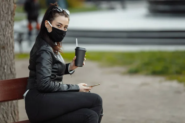 Portrait de femme brune portant un masque de protection noir assise sur un banc dans le parc, journée ensoleillée du printemps. Femme parlant sur téléphone portable — Photo