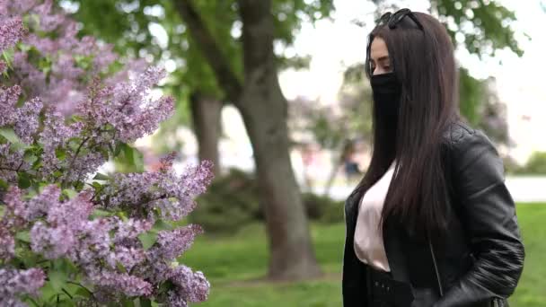 Una chica en un parque con una máscara médica negra huele flores, huele lilas en un cálido día de primavera. Día soleado de verano. Cuarentena, coronovirus. COVID 19. Alergia de primavera — Vídeos de Stock