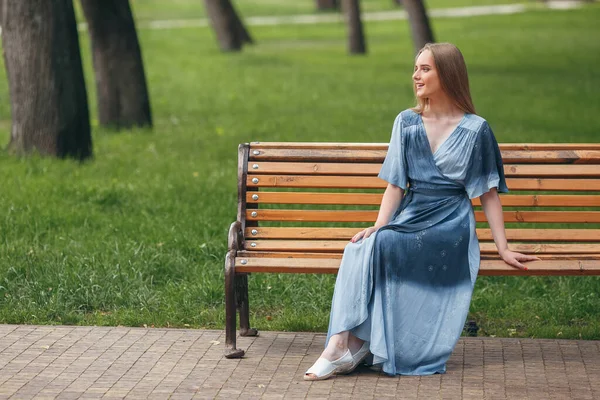 Όμορφο κορίτσι κάθεται σε ένα παγκάκι, μελαχρινή σε ένα φωτεινό φόρεμα, ηλιόλουστη μέρα — Φωτογραφία Αρχείου