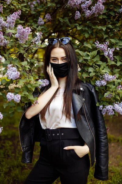 Çiçek açan leylakların arka planında tıbbi maskeli bir kız. Siyah maske.