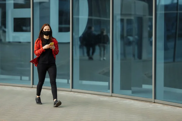 Young beautiful woman wear black face mask protection against epidemic coronavirus covid-19. Looks on phone screen. European girl medical protective mask orange business suit.