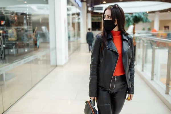 Uma menina com uma máscara preta médica está andando ao longo de um centro comercial. pandemia de coronavírus. Uma garota com uma máscara protetora está comprando no shopping — Fotografia de Stock