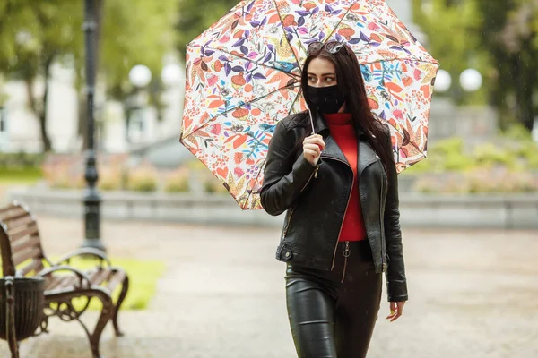 Uma menina mascarada está andando pela rua. Uma menina com uma máscara protetora caminha no parque com um guarda-chuva na chuva. Infecção pelo vírus da coronária COVID-19 — Fotografia de Stock