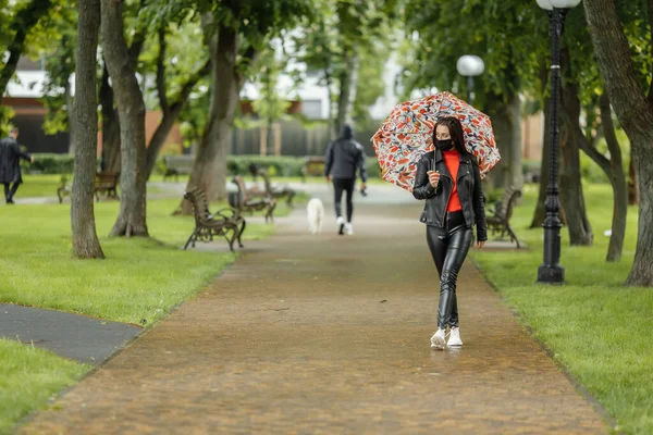 Une fille masquée marche dans la rue. Une fille portant un masque de protection se promène dans le parc avec un parapluie sous la pluie. Infection à coronavirus COVID-19 — Photo