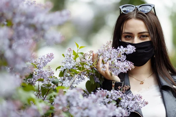Çiçek açan leylakların arka planında tıbbi maskeli bir kız. Siyah maske. Coronavirus koruması. Koronavirüs salgını. Bahar alerjisi
