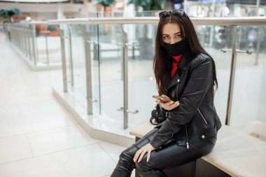 Alışveriş merkezinde siyah maskeli ve cep telefonu olan bir kız. Koronavirüs salgını. Maskeli bir kadın alışveriş merkezinde duruyor. Koruyucu maskeli bir kız alışveriş merkezinde alışveriş yapıyor.