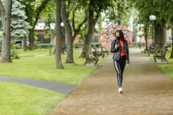 Una ragazza mascherata sta camminando lungo la strada. Una ragazza con una maschera protettiva cammina nel parco con un ombrello sotto la pioggia. Infezione da coronavirus COVID-19 — Foto Stock