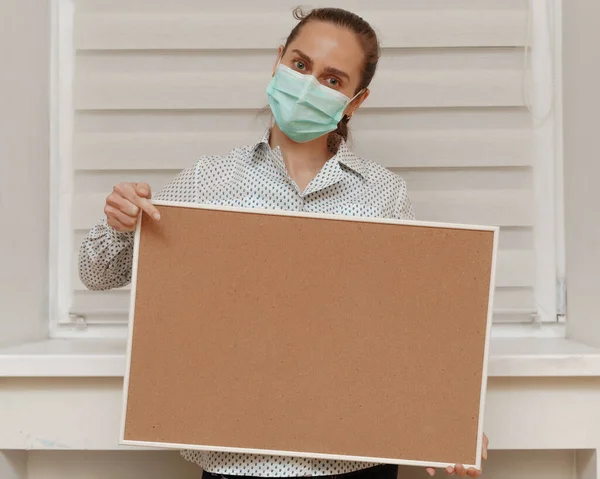 A young woman in a mask shows a finger on a blank cork board, standing on a white background. Copy space on whiteboard for image or message. Young woman. Concept of paying attention to your promise