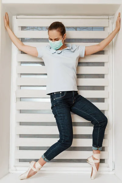 A female ballerina in a mask in casual clothess, jeans and a t-shirt in Pointe shoes at home on the windowsill posing for the camera. The concept of home occupation in quarantine