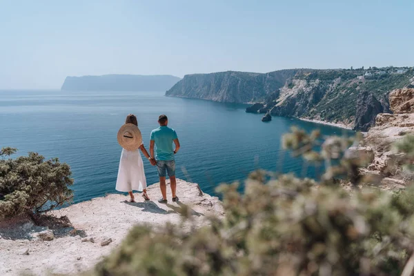 Young couple on vacation are standing with their backs holding hands looking at a beautiful seascape. In bright clothes, the girl has a hat behind her back. The concept of romance, travel, adventure
