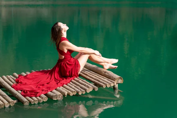 A girl with fluffy hair in a red light dress barefoot in the summer in a forest on a lake sits on a pantone on a wooden pier bridge. The concept of harmony with nature, leisure and travel, blogger