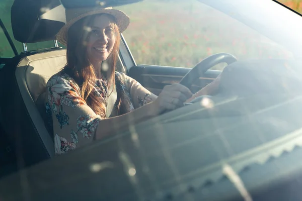 A smiling woman in a hat and colored dress sits behind the wheel of a car, behind the windshield. In the window blooming field of poppies.