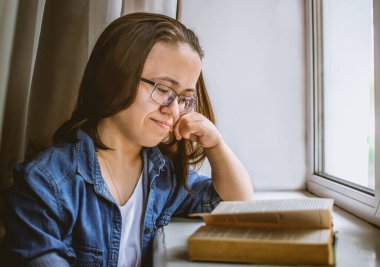 Gözlüklü kız ilginç bir kitap okuyor ve pencerenin yanında gülümsüyor.