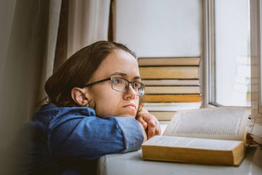 Gözlüklü kız açık bir kitabın yanında hayaller kuruyor ve pencereye bakıyor..