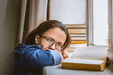 Gözlüklü bir kız pencere pervazında açık bir kitabın yanında sıkılır. Gözlüklü öğrenci evde çalışırken sıkıldı.